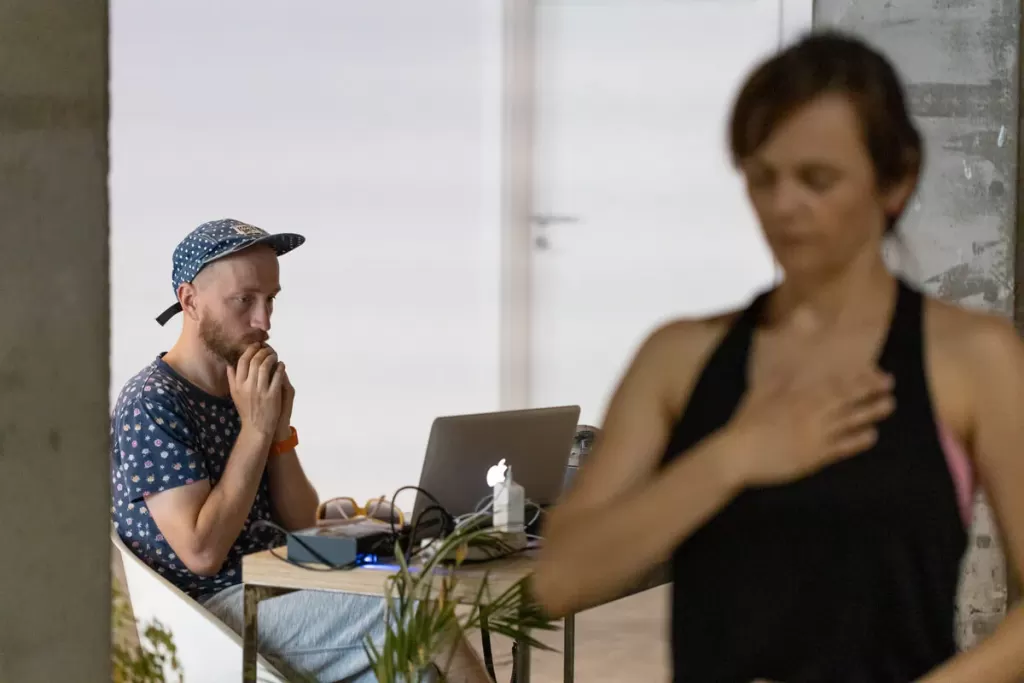 Mężczyzna siedzi przed laptopem. Kobieta stoi i trzyma rękę na klatce piersiowej.