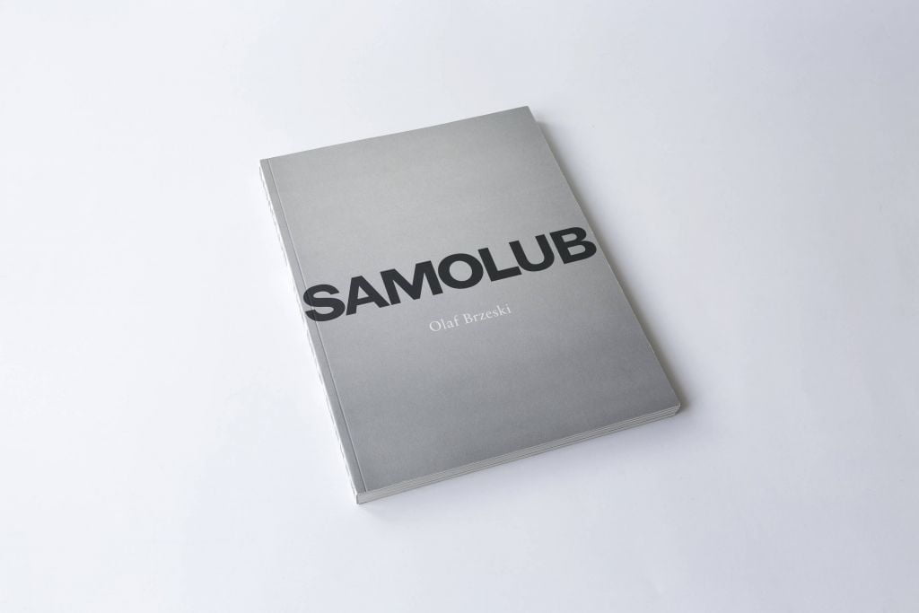 Na białym tle ksiązka o szarej okładce. Na środku czarny napis SAMOLUB, pod spodem podpis Olaf Brzeski.