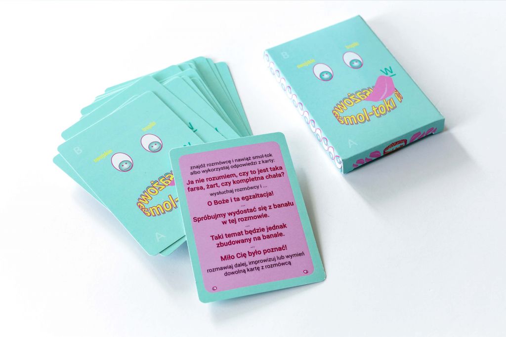 Na białym tle zestaw kart do gry – karty są w kolorze turkusowym oraz różowym. Na pudełku znajduje się rysunek uśmiechniętej buzi.