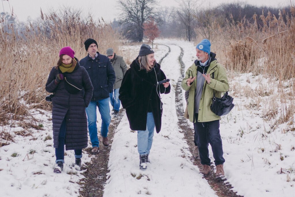 Dwóch mężczyzn i trzy kobiety idące przez zaśnieżone pole. Ziemię pokrywa śnieg, jest zima, wszyscy są ubrani w czapki i grube kurtki. 