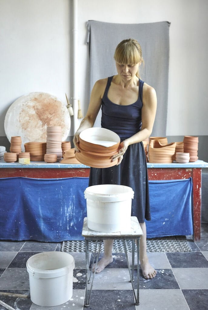 Kobieta na zdjęciu wylewa do białego wiadra masę ceramiczną. Za nią, na stole, stoją ceramiczne naczynia