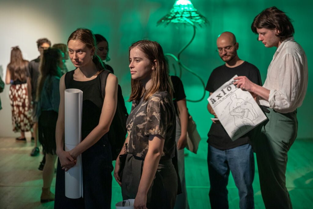 zdjęcie osób w galerii podczas wydarzenia. w tle zielone światło i prace na wystawie