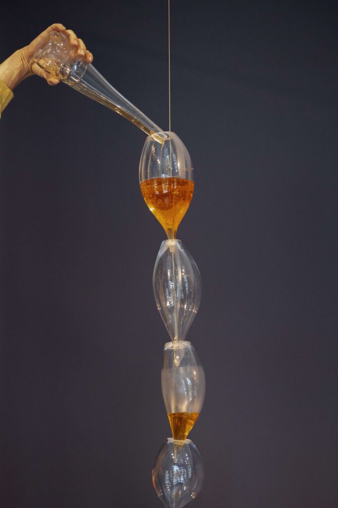 Górna część szklanej pracy na szarym tle. Praca wygląda jak lejek z połączonych ze sobą kieliszków. Po lewej stronie dłoń najewająca pomarańczową ciecz do lejka.