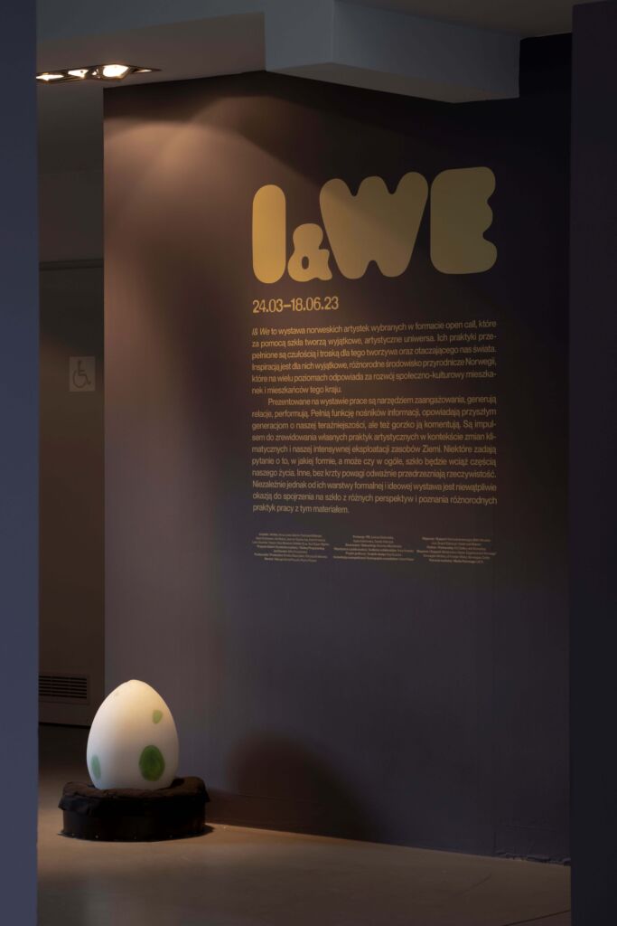 Duże, białe jajko z zielonymi kropkami w oddaleniu na tle ściany z wyklejką na temat wystawy i dużym napisem I & WE.