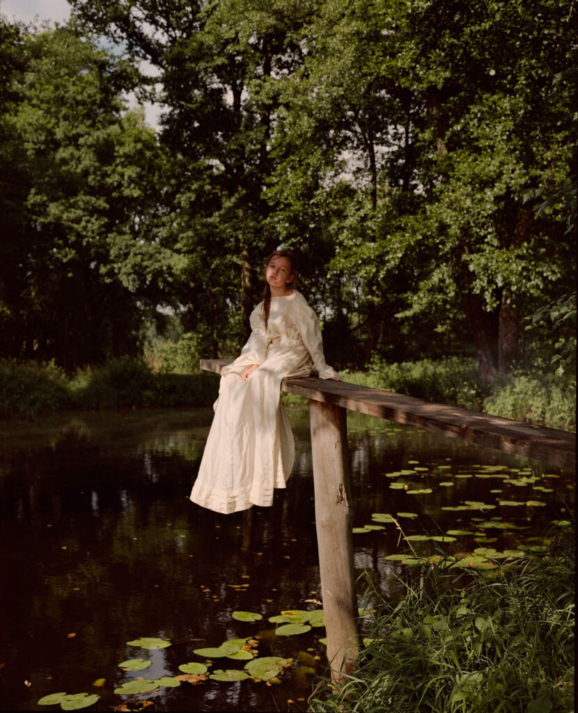 Dziewczynka w sukni ślubnej siedzi na drewnianym konarze nad wodą z nenufarami, wokół drzewa