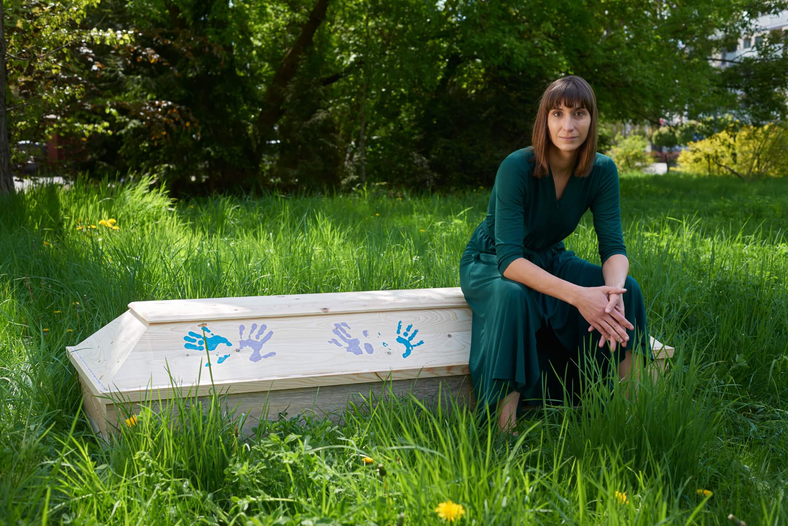 Anja Franczak siedzi na białej trumnie, na której widać kolorowe odciski dłoni. Anja ma na sobie długą, ciemnozieloną sukienkę. W tle bujna zieleń, trawa i drzewa