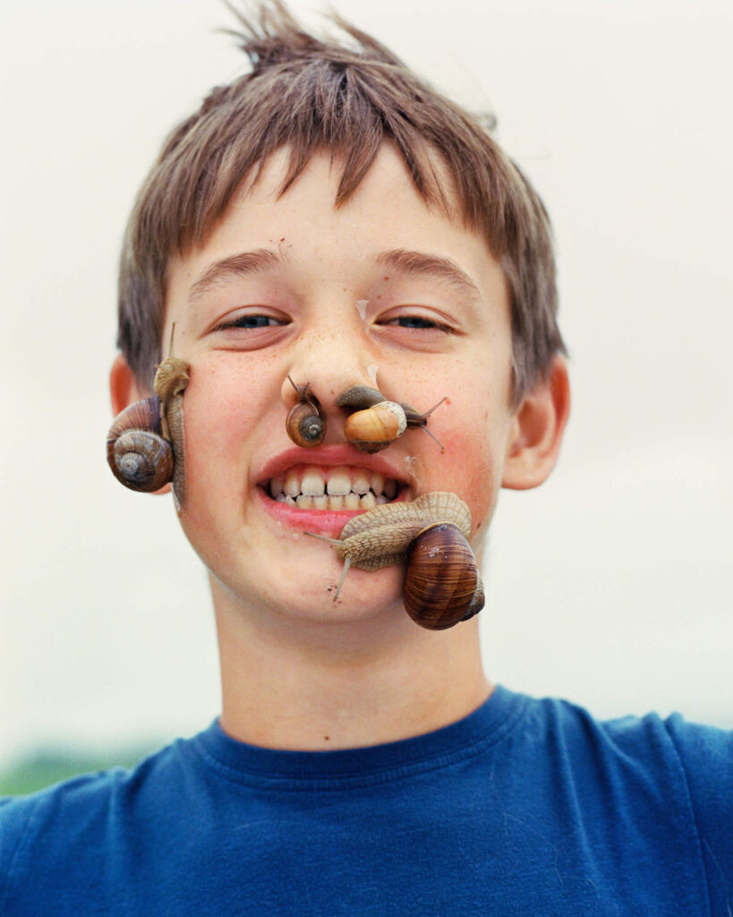 Portret młodego chłopca na jasnym tle, z ciemnymi włosami i wyszczerzonymi zębami oraz czterema ślimakami winniczkami różnej wielkości, pełzającymi po jego twarzy i wchodzącymi mu do nosa.
