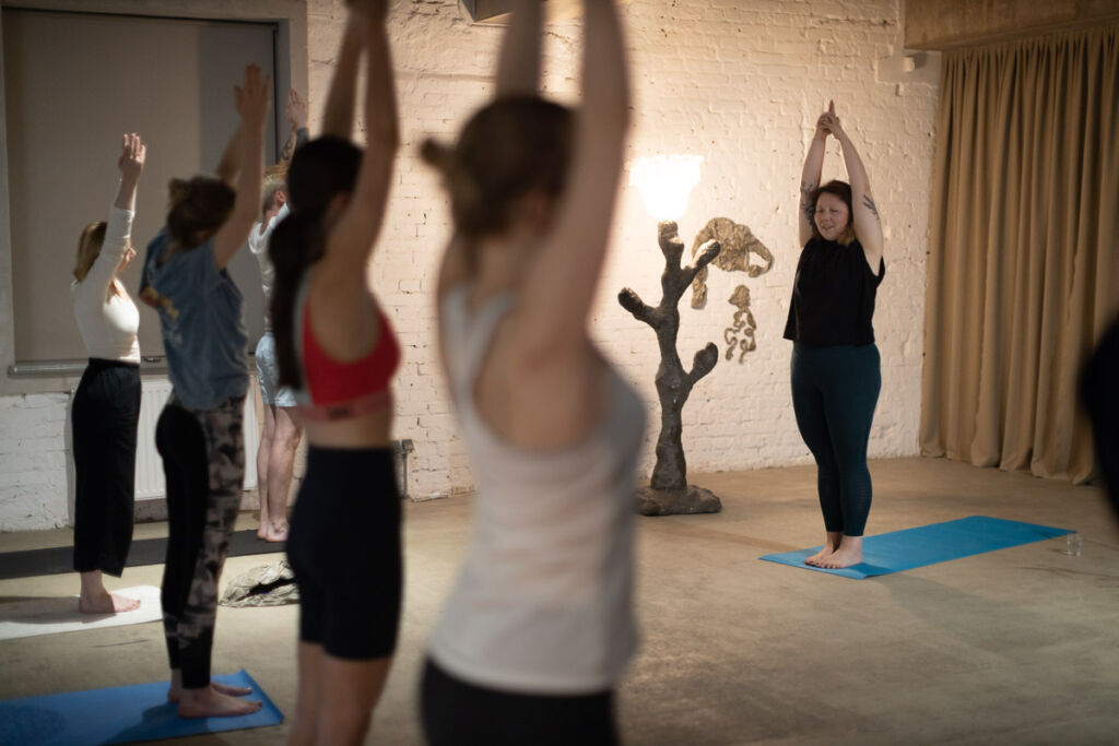 Osoby praktykujące jogę stoją w linii z podniesionymi rękoma, na przeciwko na macie prowadząca pokazuje pozycję, w przestrzeni znajdują się instalacje artystyczne.