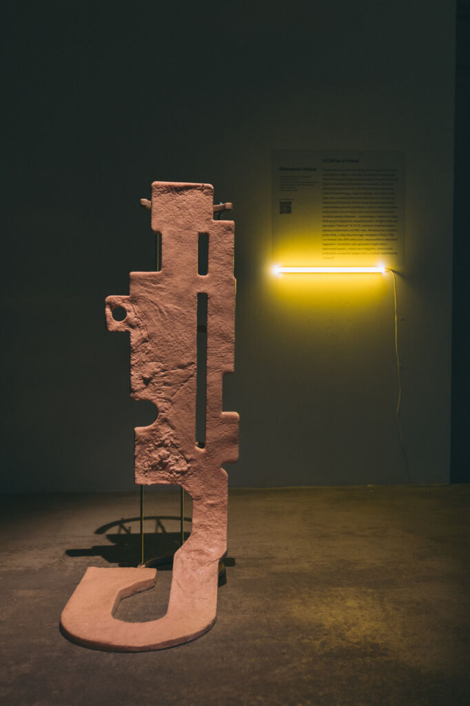 Rzeźba w kolorze różowym, podłużna, wysoka, w tle fragment opisu pracy podświetlony od dołu żółtym neonem