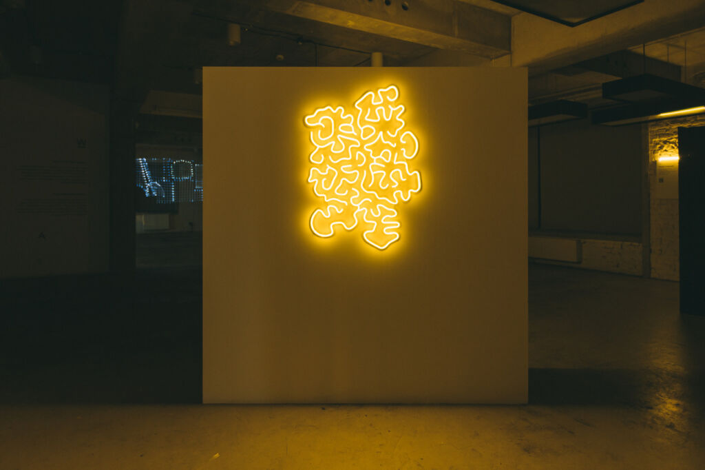 abstrakcyjny wzór przypominający podziemną grzybnię w formie neonu, powieszony na ścianie i świecący się na żółto
