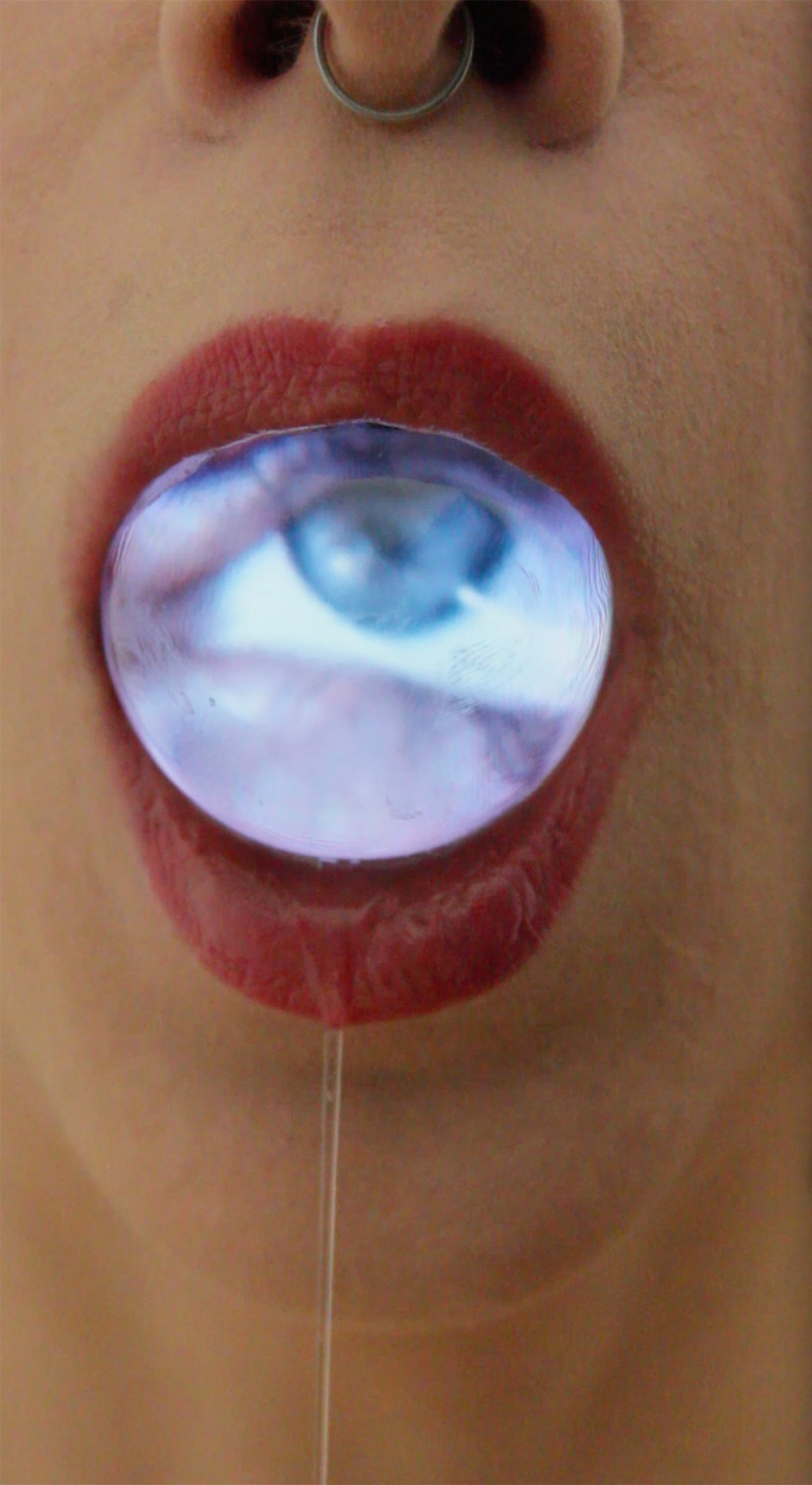 zbliżenie na usta pomalowane szminką, a w nich szklana kula o powierzchni ekranu, na której wyświetla się oko. Z ust wycieka strużka śliny.