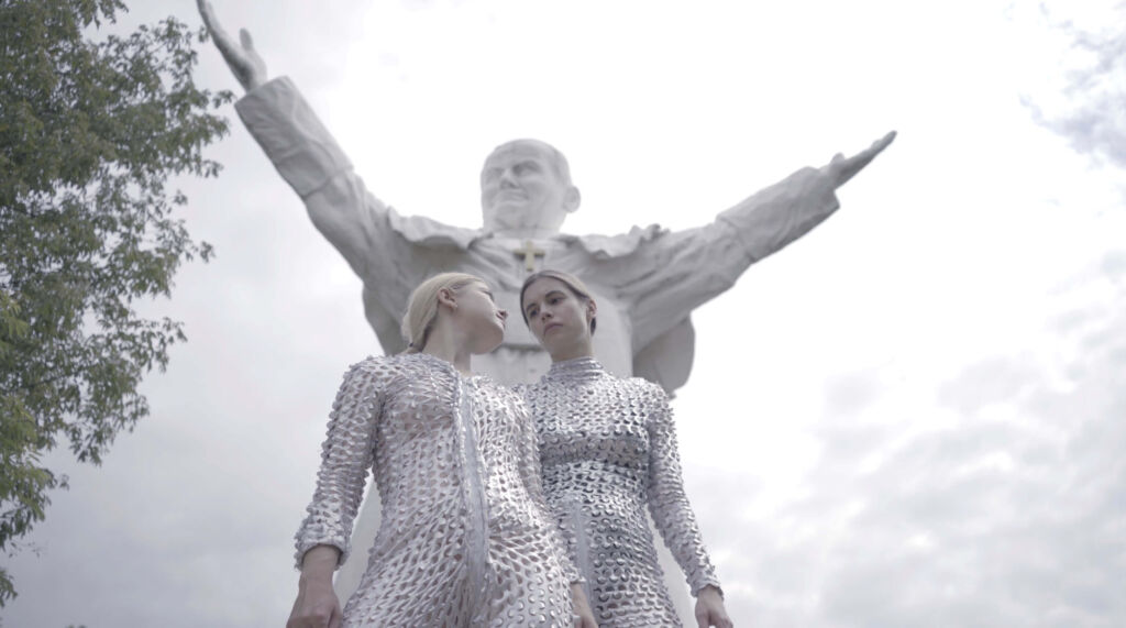 Two women in silver outfits standing next to a statue.