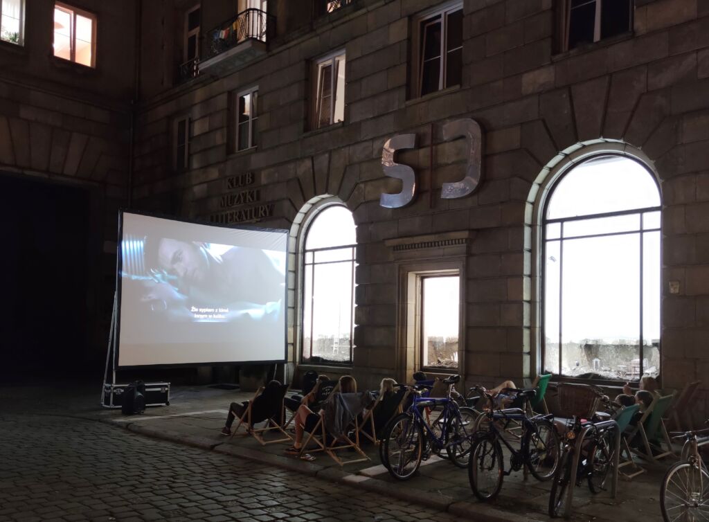 Plenerowa projekcja filmu przed galerią SIC, widzowie na leżakach i grupa rowerów zaparkowana przed budynkiem.