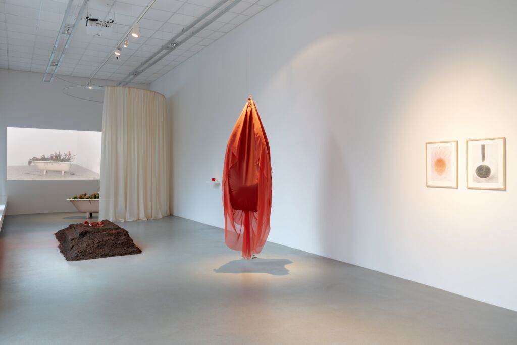 Widok wystawy z czerwonym obiektem z tkaniny, kopcem z ziemi i projekcją wideo na ścianie