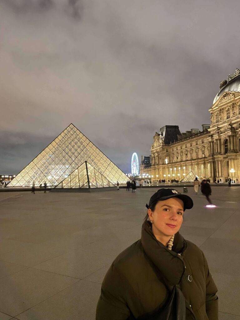 Zdjęcie Zofii Pałuchy w puchowej kurtce i czapce z daszkiem na tle muzeum Louvre w Paryżu.