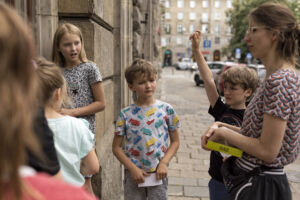 A group of children attentively listening to a woman pointing upwards during an outdoor activity.