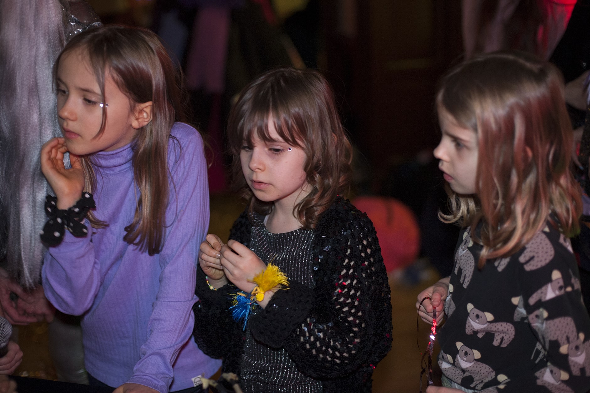 Trójka dzieci na imprezie w pomieszczeniu, uważnie przyglądająca się czemuś poza kamerą, wyrażająca ciekawość i koncentrację.