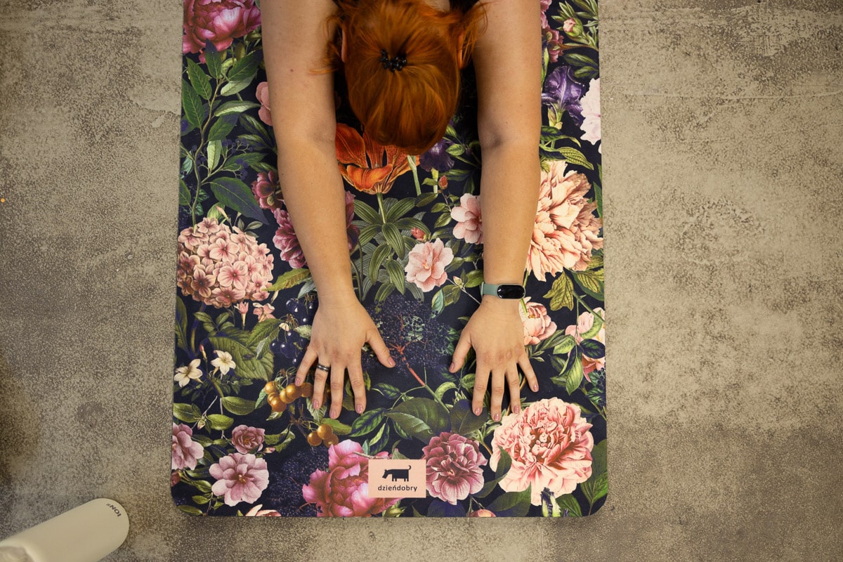 Kobieta leżąca twarzą w dół na kolorowej, kwiecistej macie do jogi, z ramionami wyciągniętymi do przodu, na ręce ma czarny zegarek.
