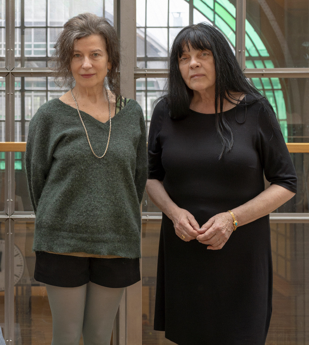 Dwie kobiety stoją w pomieszczeniu przed oknem. Jedna ma na sobie zielony sweter i czarne spodenki, a druga czarną sukienkę. Oboje mają ciemne włosy i patrzą w kamerę.