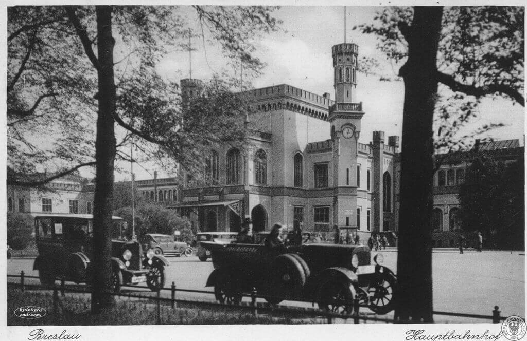 Historyczna fotografia Breslau Hauptbahnhof, przedstawiająca klasyczne samochody i pojazd zamiatający ulice przed dworcem kolejowym. Scenę otaczają drzewa, a po prawej stronie widoczna jest wieża zegarowa.