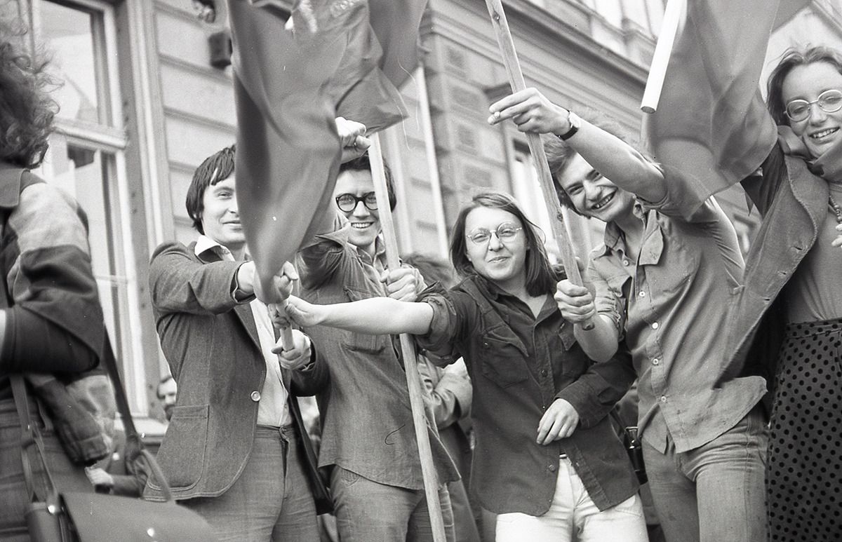 Grupa młodych ludzi trzymających flagi i uśmiechających się podczas imprezy plenerowej. Obraz jest czarno-biały.
