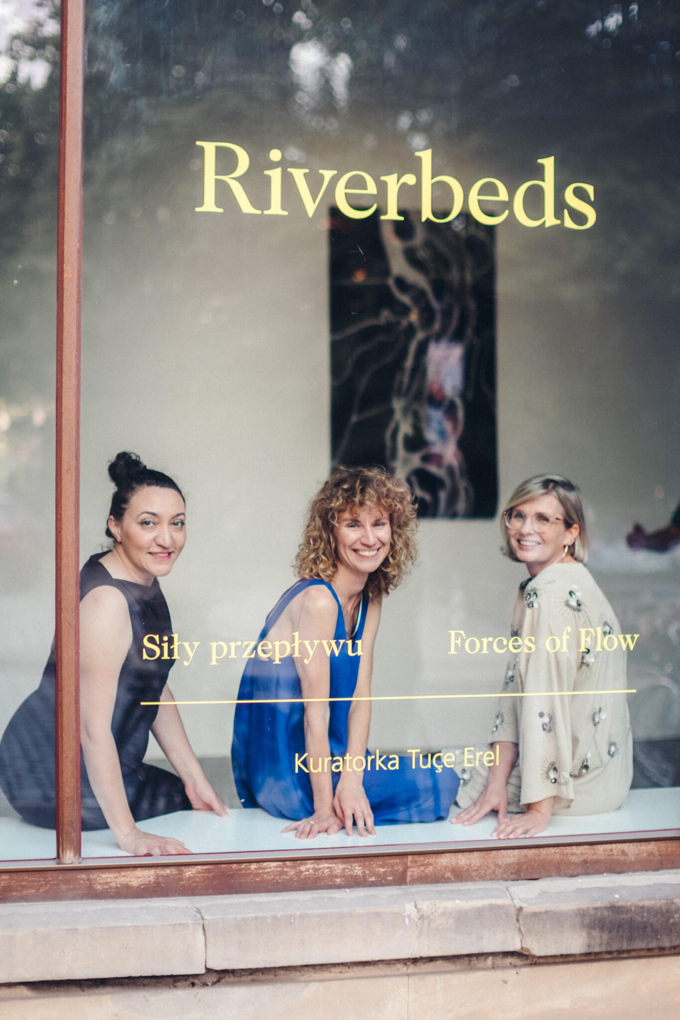 Trzy kobiety siedzą przy oknie, na którym widnieją napisy „Riverbeds” i „Forces of Flow” w języku angielskim i polskim oraz „Kuratorka Tuçe Erel”. uśmiechają się i stoją przed kamerą.