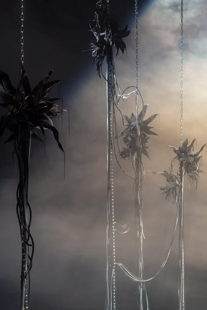 Metal abstract sculptures resembling plants, suspended on chains in a misty and dimly lit environment.