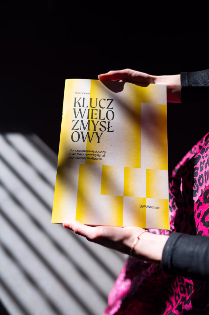 Osoba trzyma książkę zatytułowaną „Klucz Wielozmysłowy” z żółto-białym geometrycznym wzorem na okładce, oświetloną światłem słonecznym.