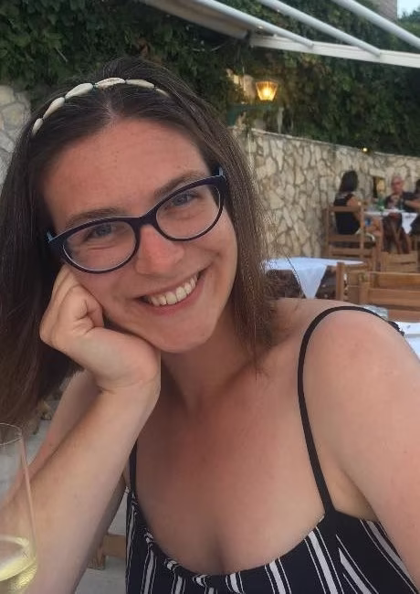 Kobieta w okularach i pasiastej sukience siedzi przy stoliku w restauracji na świeżym powietrzu, uśmiecha się do kamery, opierając głowę na ręce.
