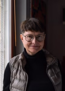 Osoba o krótkich brązowych włosach, w okularach i w kamizelce puchowej stoi w pomieszczeniu przy oknie, patrząc w kamerę z neutralnym wyrazem twarzy.