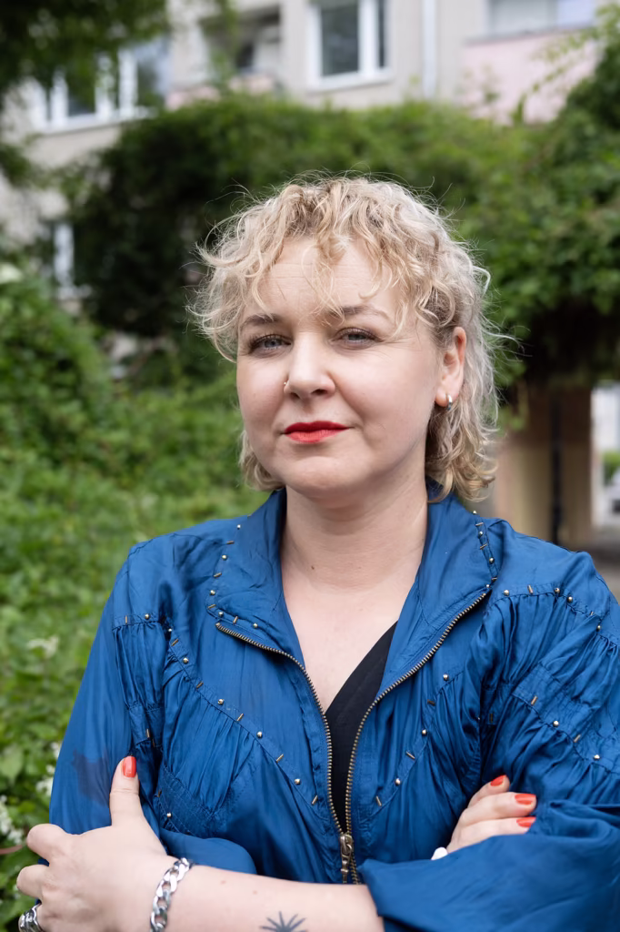 Kobieta z krótkimi, kręconymi blond włosami i czerwoną szminką stoi na zewnątrz ze skrzyżowanymi ramionami, ubrana w niebieską kurtkę i srebrną bransoletkę, na tle zieleni i budynku.