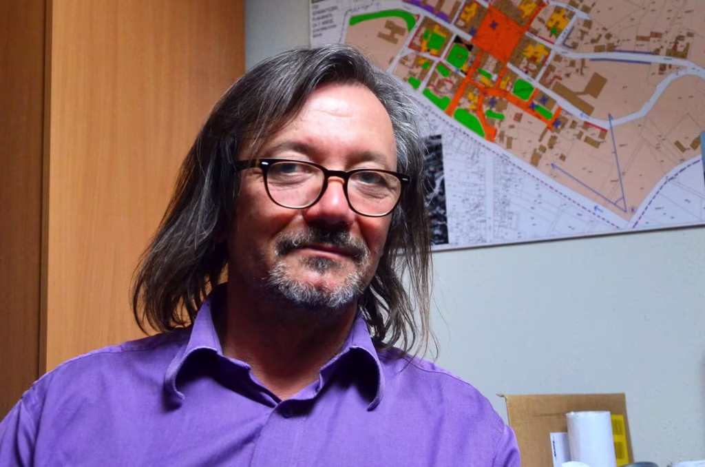Portret mężczyzny w okularach, fioletowej koszuli, włosach do ramion, w tle mapa.