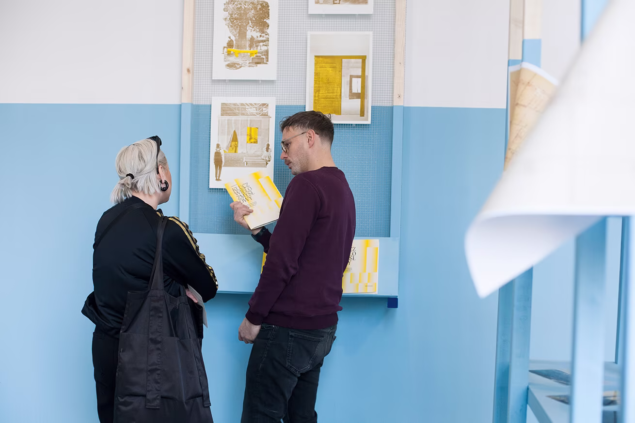Dwie osoby stoją przed niebieską ścianą, dyskutując i wskazując na rysunki architektoniczne wyświetlane w ramkach.
