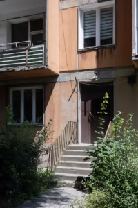 Betonowe schody z metalową poręczą prowadzą do brązowych drzwi pomarańczowego budynku mieszkalnego, otoczonego zarośniętymi zielonymi roślinami.