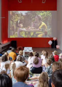 Grupa dzieci siedzi na podłodze i ogląda film animowany wyświetlany na ekranie w jasno oświetlonym pokoju udekorowanym balonami.