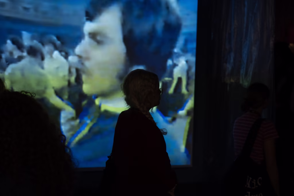 Osoba stoi w sylwetce przed dużym ekranem wyświetlającym rozmyty, zabytkowy film przedstawiający ludzi w zatłoczonym otoczeniu.