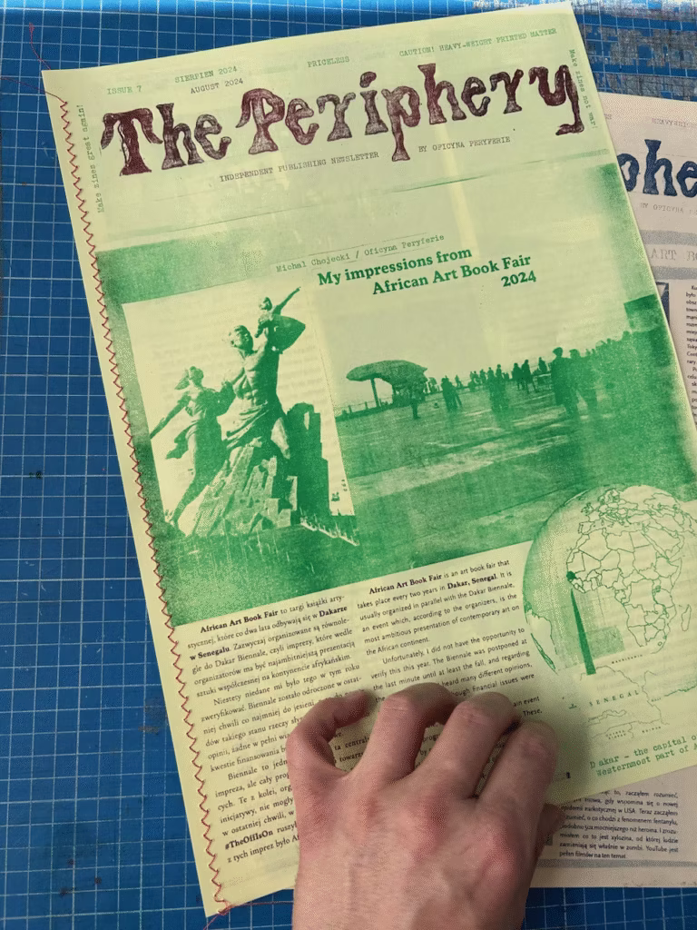 Ręka trzyma wydanie gazety "The Periphery" z artykułem zatytułowanym "Moje wrażenia z African Art Book Fair 2024" i zielonym zdjęciem posągu.