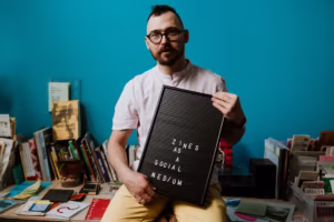 Osoba siedzi przed niebieską ścianą, trzymając tablicę z napisem "ZINES AS A SOCIAL MEDIUM", otoczoną książkami i czasopismami.
