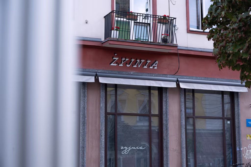 Fasada budynku z balkonem, roślinami doniczkowymi i dużymi oknami przedstawia napis "ŻYWNIA". Drzewo częściowo otacza prawą stronę obrazu.