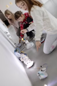 Trójka dzieci przykucnęła na szarej podłodze, wchodząc w interakcję z elektronicznymi i podświetlanymi obiektami ustawionymi w linii przy białej ścianie.