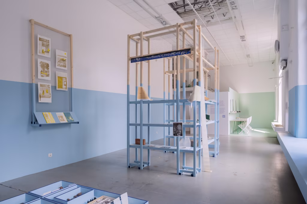 Minimalistyczna przestrzeń wystawiennicza z jasnoniebieskimi i białymi ścianami, otwartymi konstrukcjami regałów, stołami ekspozycyjnymi i oprawionymi wydrukami na ścianie.