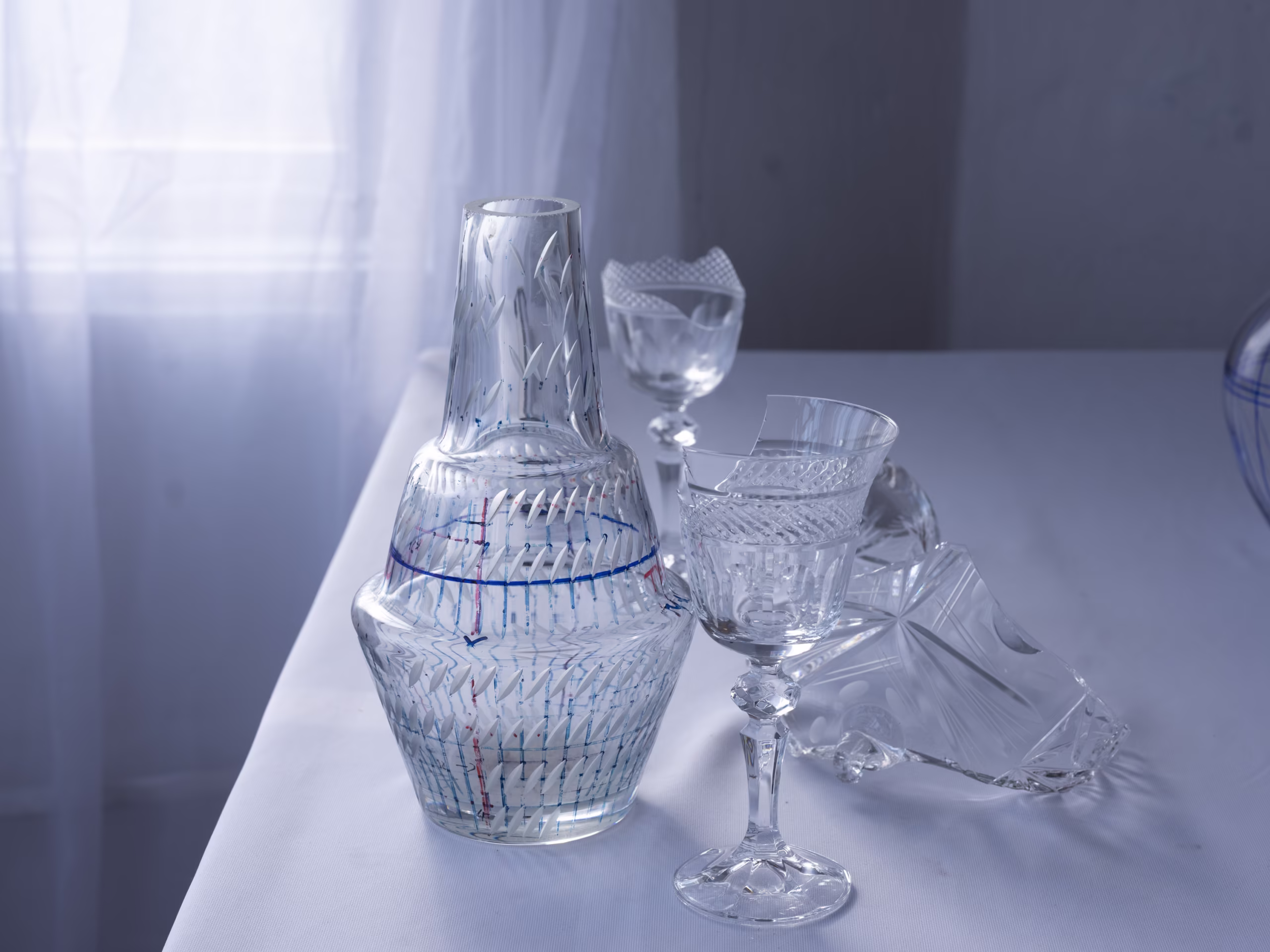 Karafka z przezroczystego, wzorzystego szkła, kilka pucharków z wytrawionego szkła i kawałek potłuczonego szkła stoją na białym stole w pobliżu zasłoniętego okna.