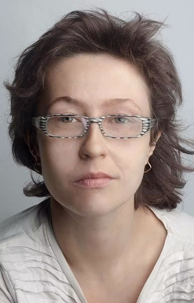 Kobieta o krótkich brązowych włosach, nosząca wzorzyste okulary, małe kolczyki kółka i jasny top, patrzy bezpośrednio w kamerę z neutralnym wyrazem twarzy.