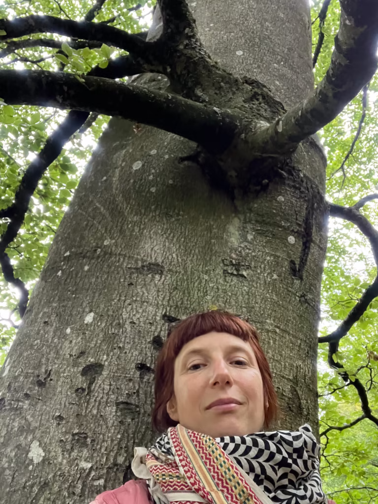 Osoba z krótkimi rudobrązowymi włosami i wzorzystym szalikiem stoi przed dużym drzewem, z zielonymi liśćmi i gałęziami widocznymi powyżej.