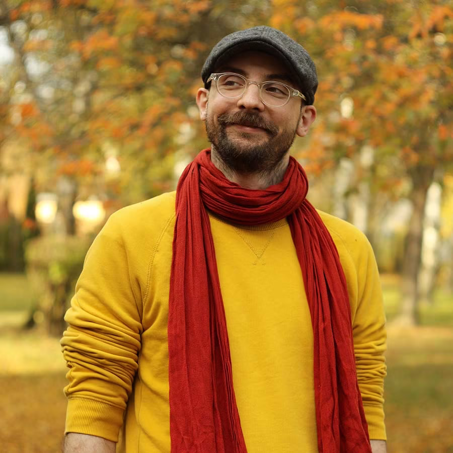 Mężczyzna w żółtym swetrze, czerwonym szaliku i szarej czapce stoi na zewnątrz z jesiennymi drzewami w tle.