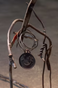 Metalowa rzeźba wykonana z wygiętych prętów i drutów, z okrągłym czarnym wisiorkiem z uśmiechniętą buźką zawieszoną w centrum.