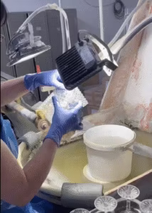 Osoba nosząca niebieskie rękawice sprawdza lub czyści przecięty szklany przedmiot pod lampą roboczą na stanowisku pracy z wodą i szklanymi naczyniami.