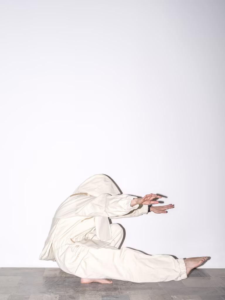 Osoba w białym stroju z kapturem przykuca blisko ziemi z rękami wyciągniętymi do przodu na tle białej ściany.