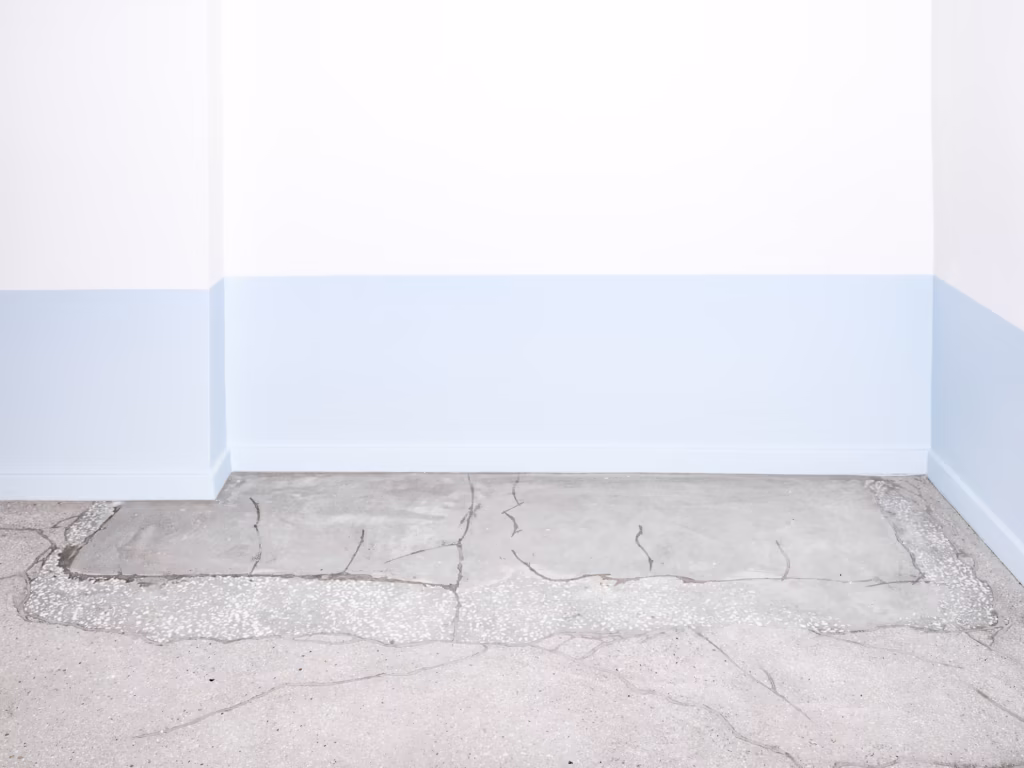 Pusty pokój z białymi ścianami, niebieską dolną krawędzią i popękaną betonową podłogą. Nie widać żadnych mebli ani dekoracji.