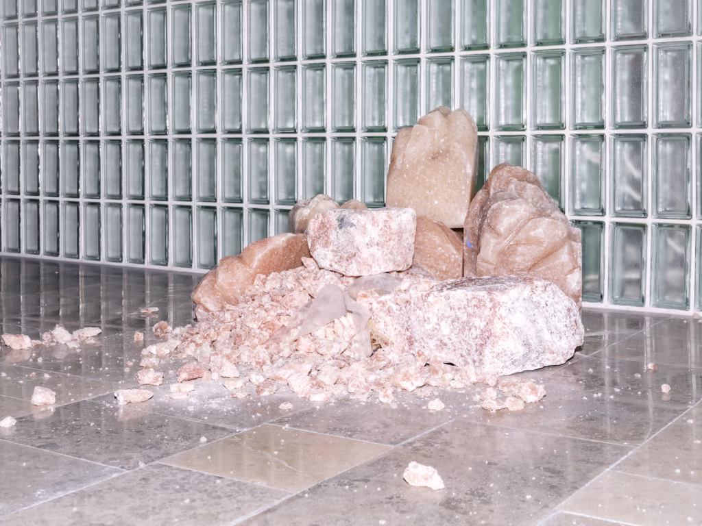 Stos dużych i małych różowawych skał leży na wyłożonej kafelkami podłodze przed ścianą wykonaną ze szklanych bloków.